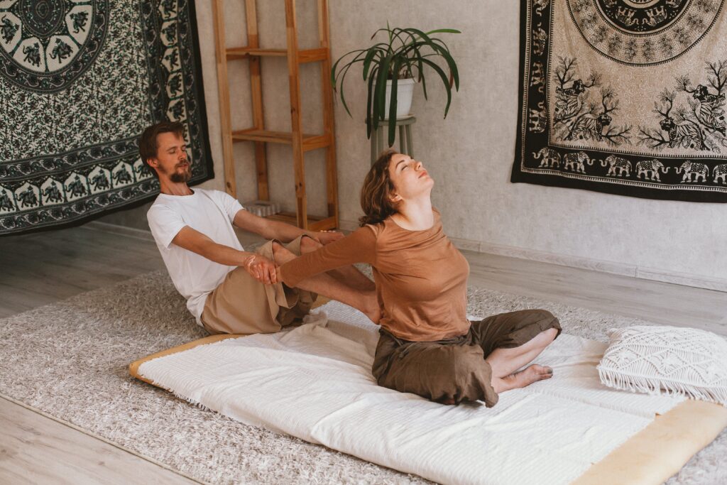 A Thai massage session in progress with a man assisting a woman in a stretching pose on a mat in a peaceful room decorated with traditional tapestries and a potted plant